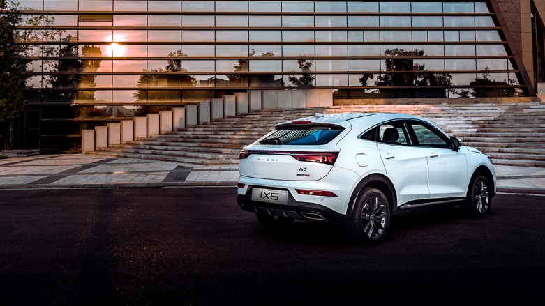 Vista trasera de SUV deportiva Glory IX5 color blanca estacionada en asfalto con un edificio moderno de fondo.